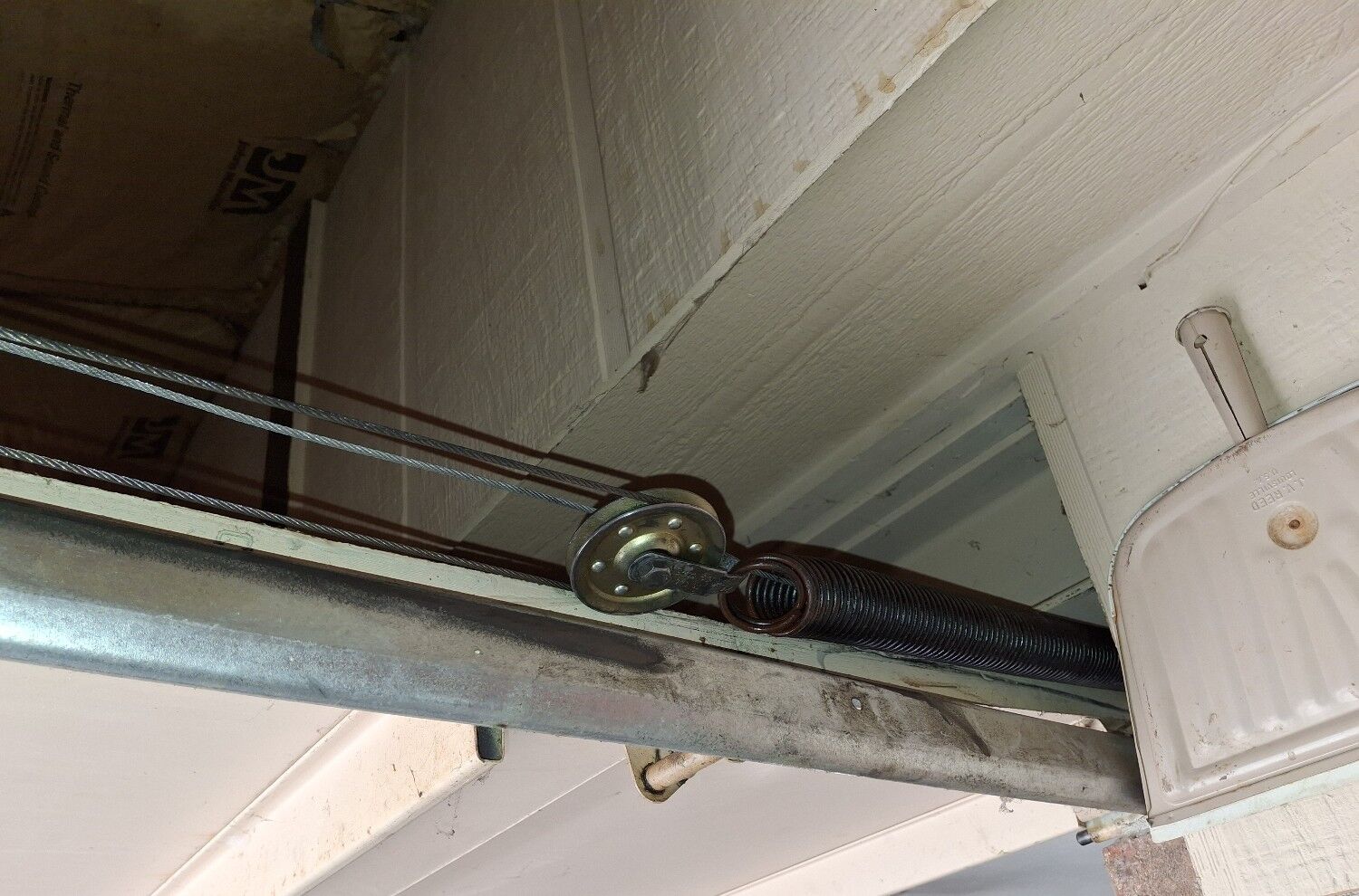 Close-up of a broken garage door extension spring stretched along the horizontal track, with visible separation in the coil and attached safety cable.