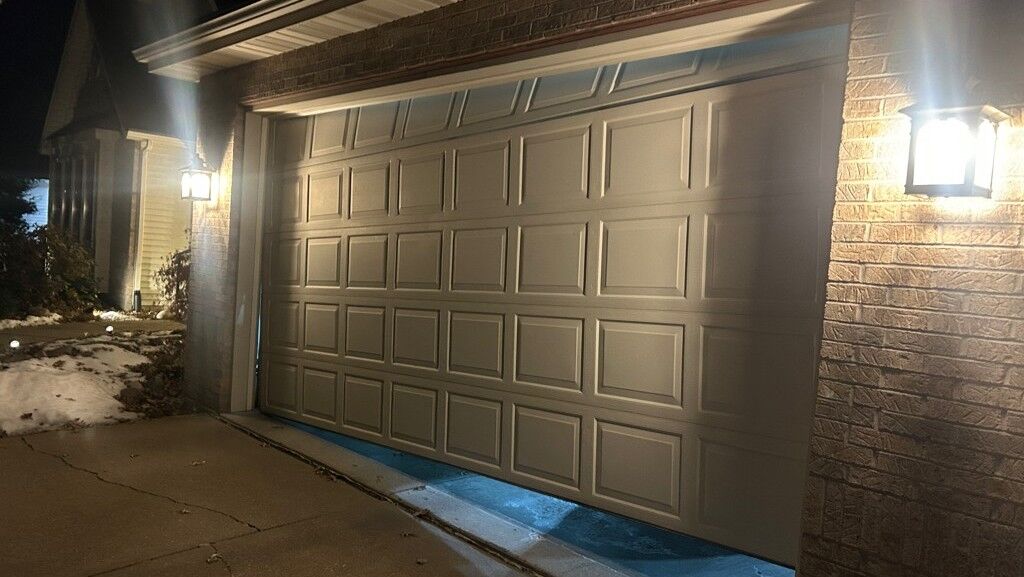 Garage door sitting unevenly with one side lower than the other, leaving a visible gap along the bottom edge while partially open at night.